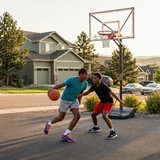 Two young men playing basketball on a driveway in a suburban neighborhood.